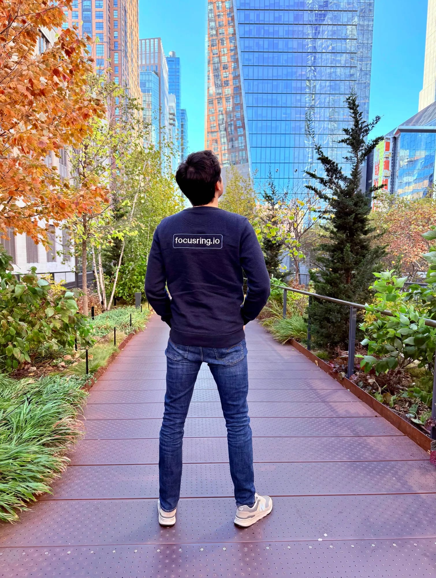 Een persoon in een focusring.io-trui, van achteren gezien, op het wandelpad van de High Line in New York, tussen planten en hoge gebouwen.